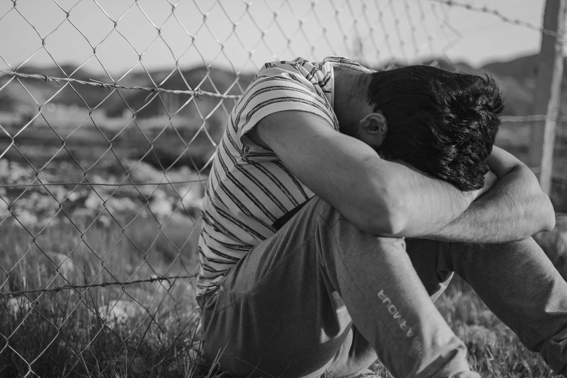 Black & white picture of a man sitting near fence while is hands are locked in on his knees.